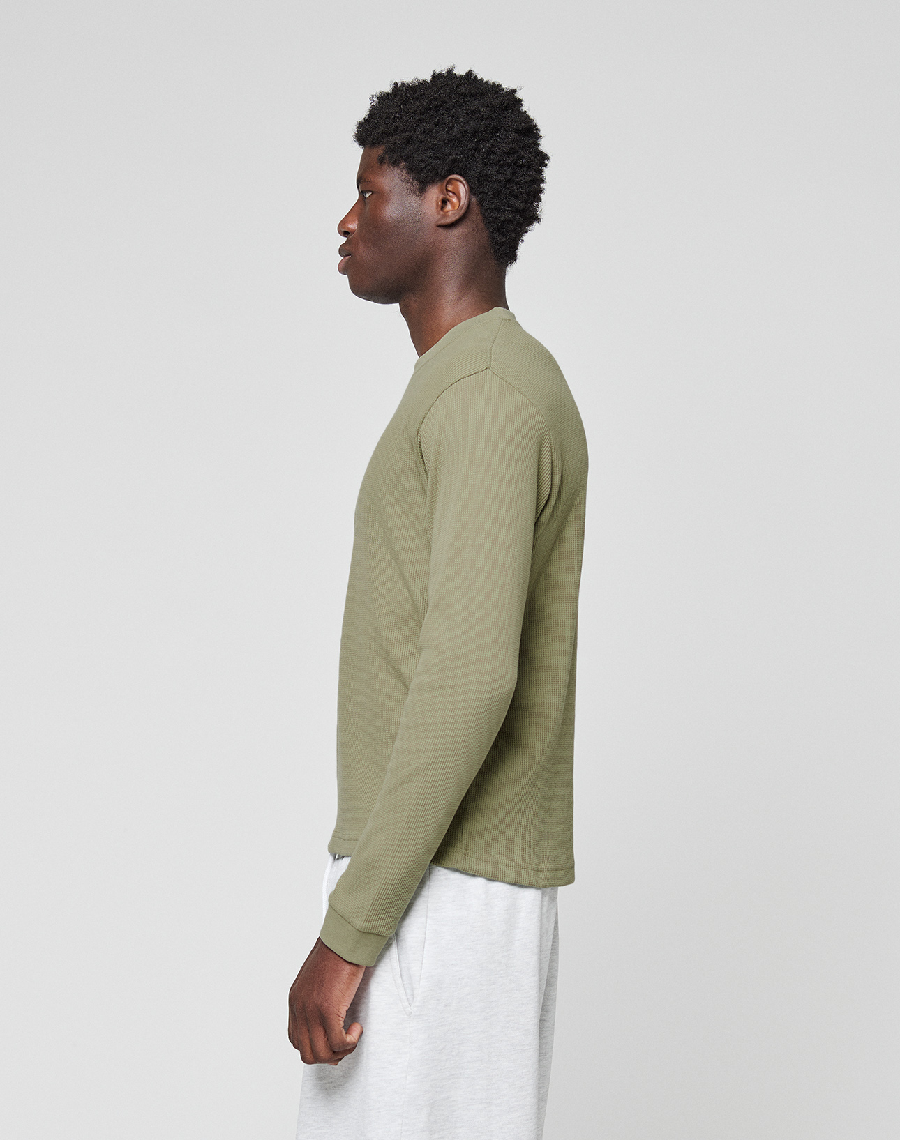 A man with short curly hair stands in profile against a plain background, wearing an olive green Basic Waffle Longsleeve by LIVE FAST DIE YOUNG and light gray shorts.
