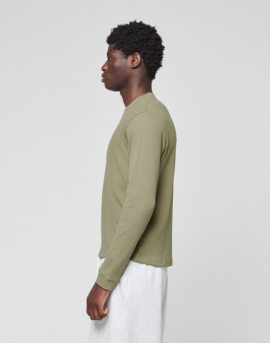 A man with short curly hair stands in profile against a plain background, wearing an olive green Basic Waffle Longsleeve by LIVE FAST DIE YOUNG and light gray shorts.