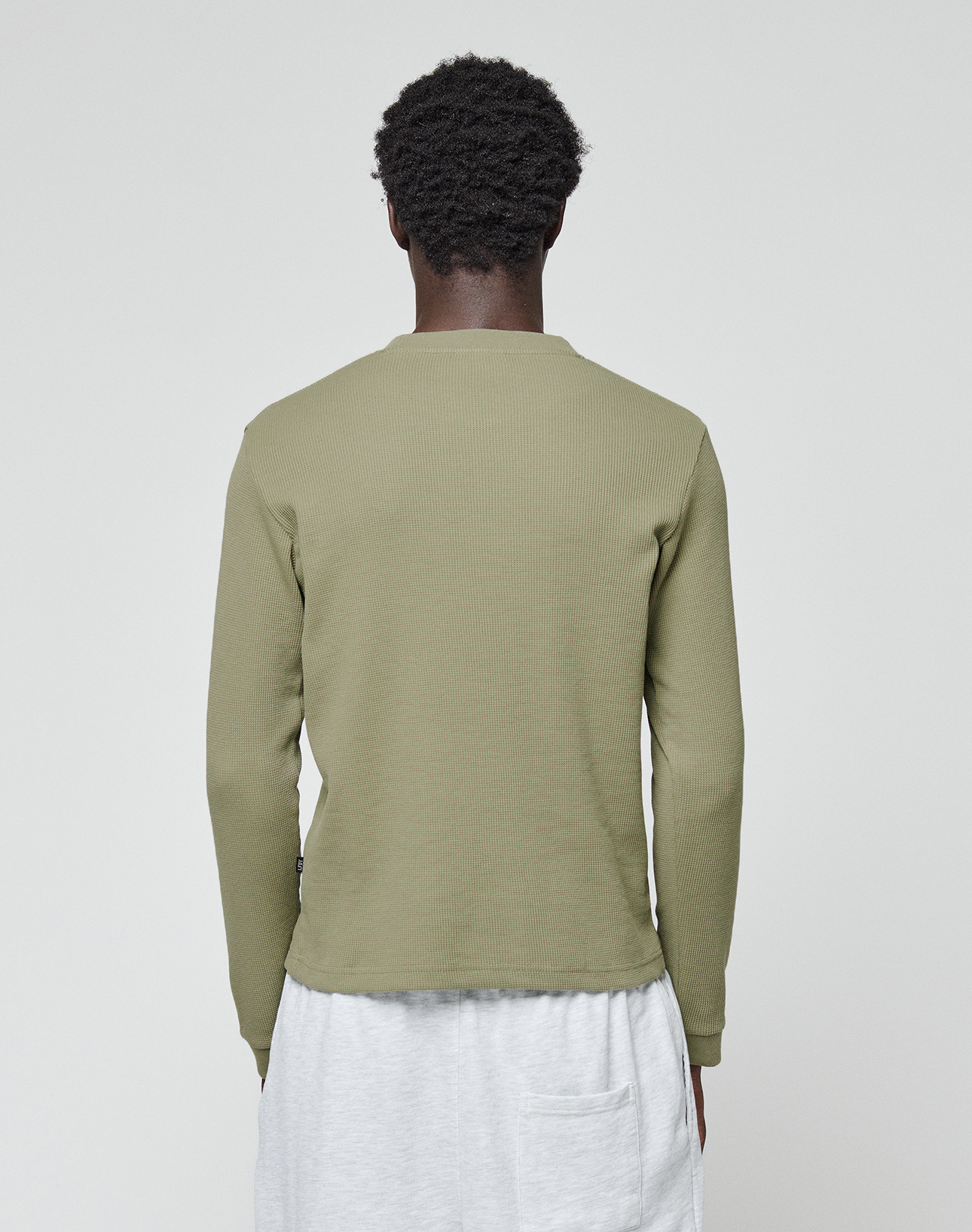A person with short curly hair stands with their back to the camera, wearing a LIVE FAST DIE YOUNG Basic Waffle Longsleeve in olive green and light gray pants with a visible back pocket, set against a plain white background.