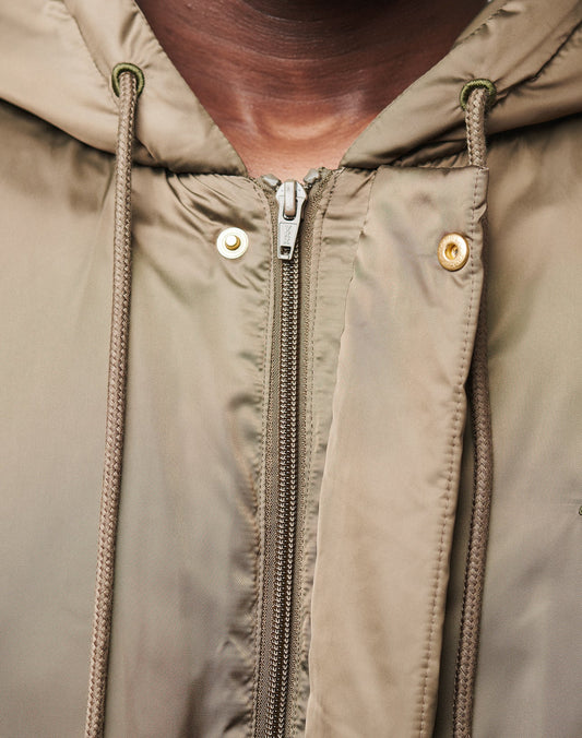 Close-up of a person in the LFDY Winter Bomber Jacket by LIVE FAST DIE YOUNG, featuring an oversize fit, brownish-green color, hood with drawstrings, zipper and metallic buttons. The chin and neck appear above the logo-stitch detail.