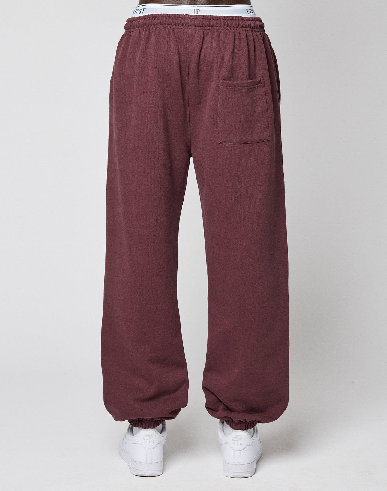 A person wears LIVE FAST DIE YOUNG Wide Cuff Jogger sweatpants in maroon, made from 100% Baumwolle French Terry, with a front pocket and white waistband, paired with white sneakers against a plain light background.