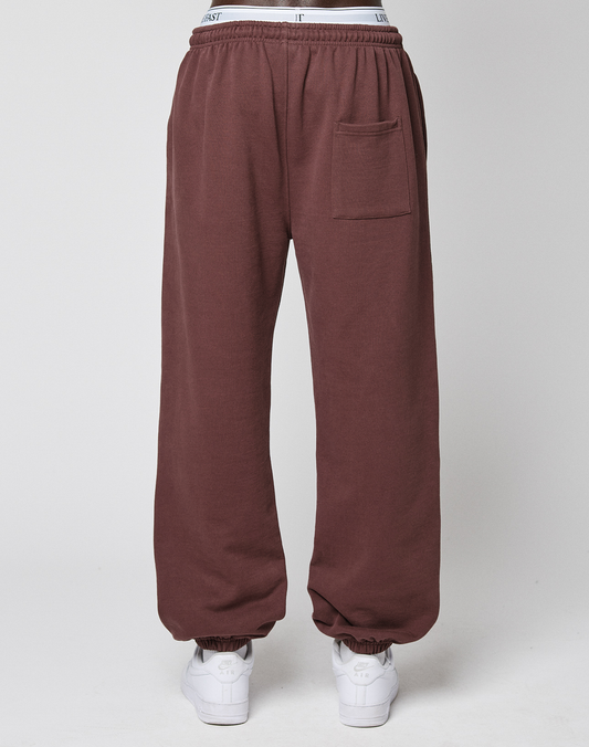 A person wears loose-fitting brown Wide Cuff Jogger by LIVE FAST DIE YOUNG, made from soft French Terry with an elastic waistband and side patch pocket, styled with white sneakers. Photo is taken from behind against a plain light background.