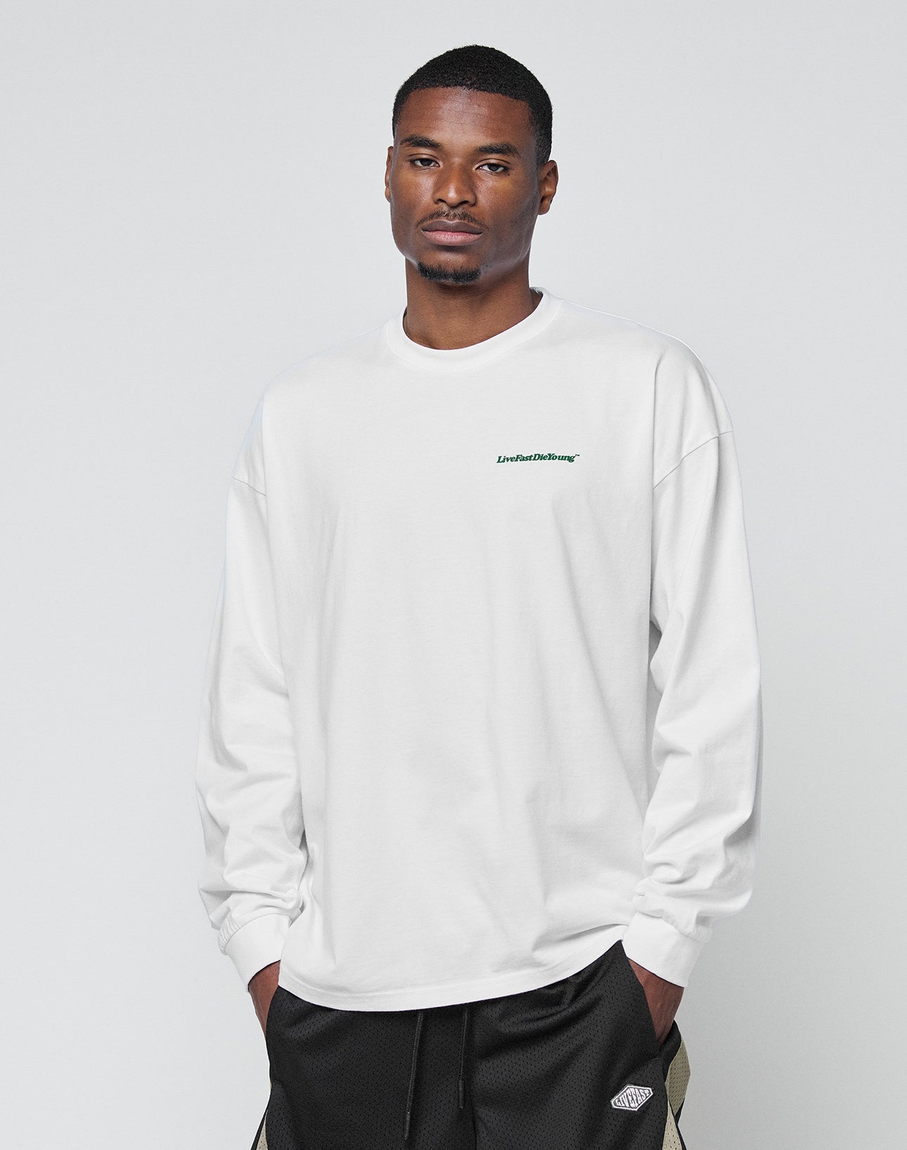 A man wears the LIVE FAST DIE YOUNG Basic Longsleeve in white, featuring a small green chest logo, paired with black athletic shorts, standing against a plain light gray background.