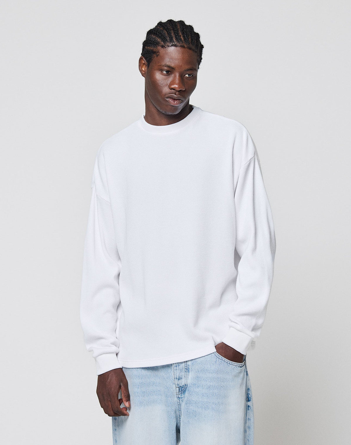 A man with braided hair wears the LIVE FAST DIE YOUNG Blank Waffle Longsleeve aus 100% Baumwolle and light blue jeans, standing against a plain light background with his hands relaxed by his sides.