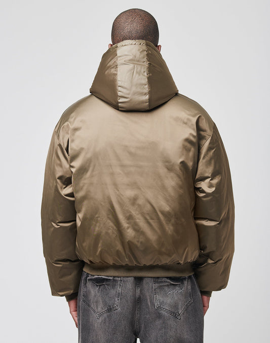 A person faces away, wearing the LFDY Winter Bomber Jacket by LIVE FAST DIE YOUNG—a shiny olive green hooded puffer with drawstring hood—and faded black oversized jeans, set against a plain light gray background.