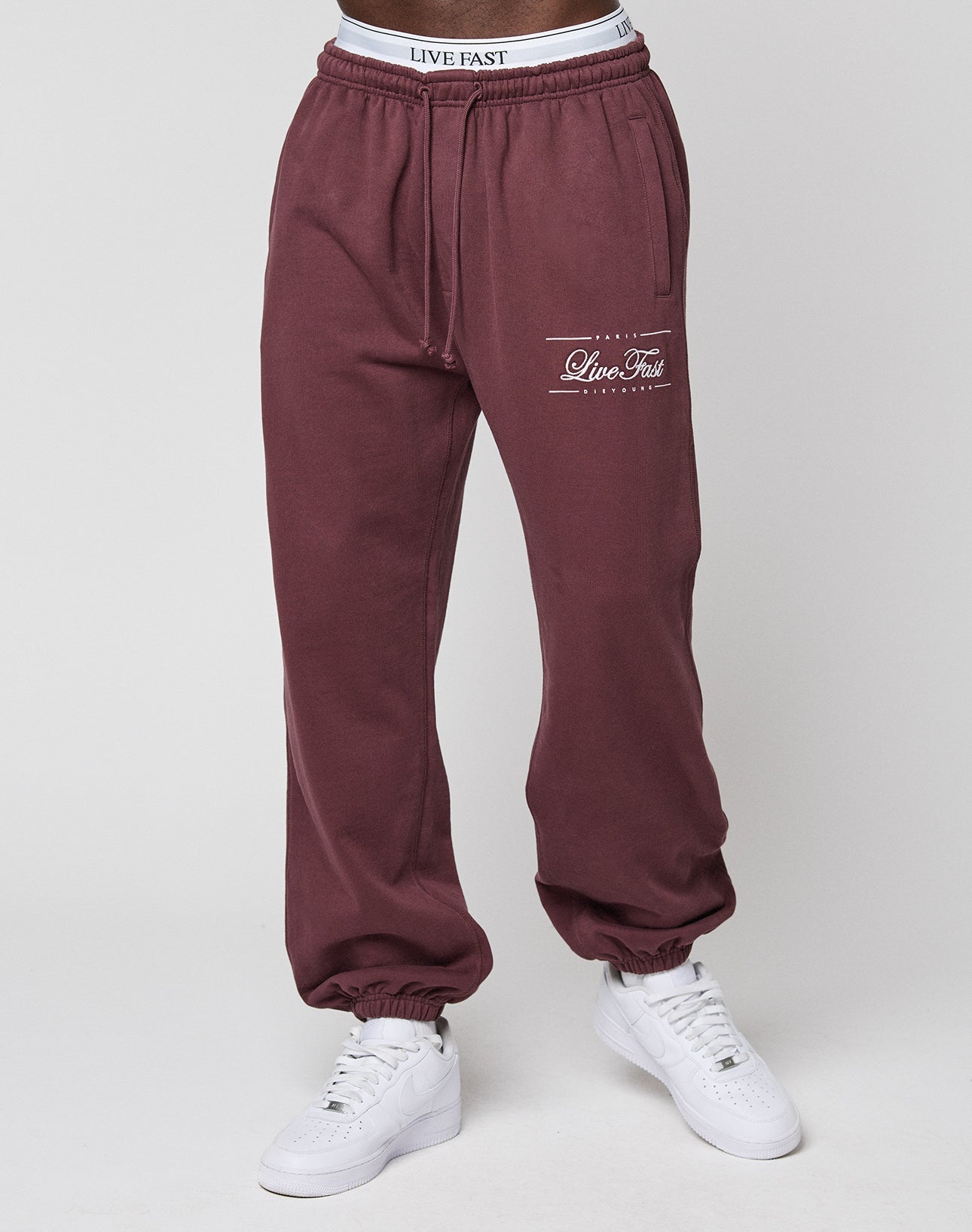 A person in maroon Wide Cuff Jogger sweatpants by LIVE FAST DIE YOUNG, featuring "Live Fast" text on the left thigh and a branded waistband, paired with white sneakers against a plain neutral background.