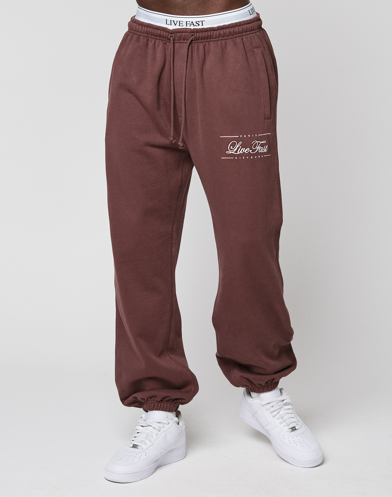 Person wears maroon Wide Cuff Jogger by LIVE FAST DIE YOUNG, featuring the Live Fast logo on the left thigh, a white waistband visible above, and white sneakers, standing against a plain light background.