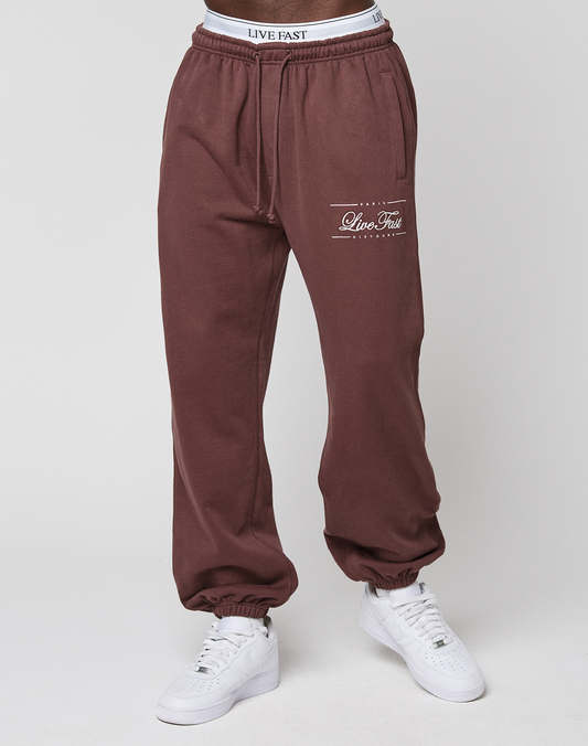 Person wears maroon Wide Cuff Jogger by LIVE FAST DIE YOUNG, featuring the Live Fast logo on the left thigh, a white waistband visible above, and white sneakers, standing against a plain light background.