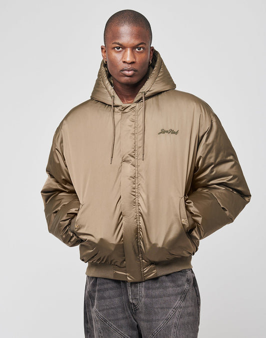 A person in the LIVE FAST DIE YOUNG LFDY Winter Bomber Jacket—an oversize light brown puffer with Kapuze mit Kordeln—and dark gray pants stands against a plain light gray background, hands in pockets and looking at the camera.