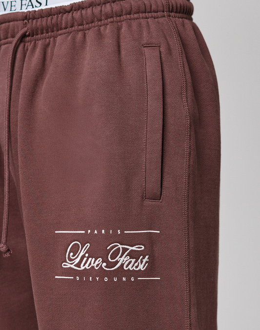 Close-up of maroon LIVE FAST DIE YOUNG Wide Cuff Jogger with elastic waistband and side pocket, showcasing white "Paris Live Fast Die Young" text near the upper thigh. The waistband and part of a matching shirt are also visible.