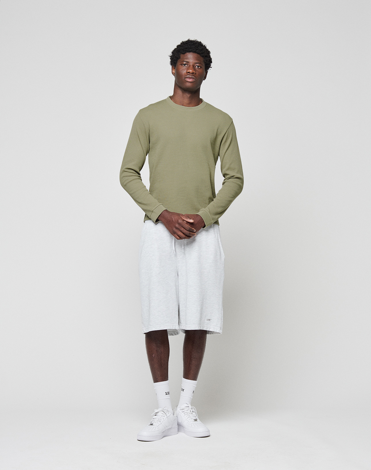 A man stands against a white background wearing the LIVE FAST DIE YOUNG Basic Waffle Longsleeve in light green, light gray knee-length shorts, white socks, and sneakers. He faces forward with hands clasped; Flag-Label detail included.