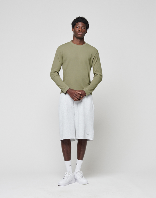 A man stands against a white background wearing the LIVE FAST DIE YOUNG Basic Waffle Longsleeve in light green, light gray knee-length shorts, white socks, and sneakers. He faces forward with hands clasped; Flag-Label detail included.