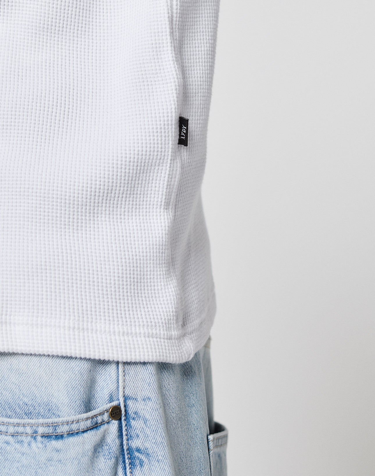 Close-up of a person wearing the LIVE FAST DIE YOUNG Blank Waffle Longsleeve, an oversized textured white shirt made from 100% cotton, featuring a small black tag, paired with light blue denim jeans. Focus is on the shirt’s hem and waistband.