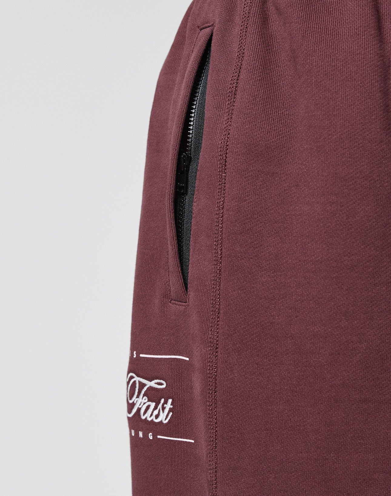 Close-up of maroon LIVE FAST DIE YOUNG Wide Cuff Jogger in 100% Baumwolle, showing a vertical zippered pocket and part of a white cursive "Fast" logo. Features a wide fit against a plain light gray background.