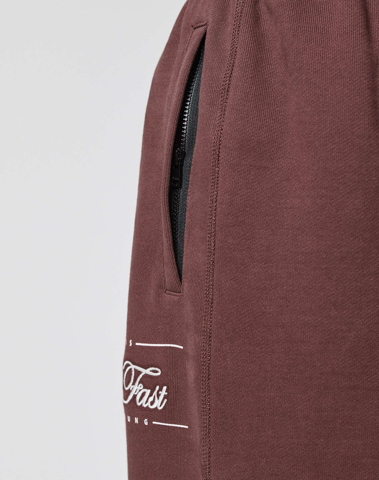 Close-up of a brown Wide Cuff Jogger from LIVE FAST DIE YOUNG, showing a black zippered pocket, partial white "Fast" text, soft French Terry-like fabric, visible seam, and elastic waistband.