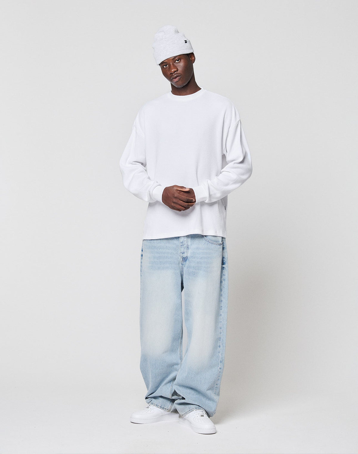 A person in a LIVE FAST DIE YOUNG Blank Waffle Longsleeve, white beanie, light blue baggy jeans, and white sneakers stands against a plain white background with hands clasped in front.