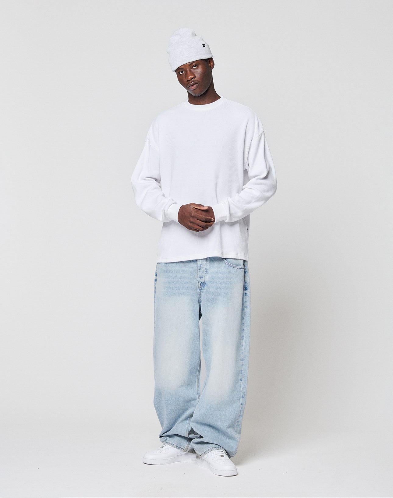 A person in a LIVE FAST DIE YOUNG Blank Waffle Longsleeve, white beanie, light blue baggy jeans, and white sneakers stands against a plain white background with hands clasped in front.