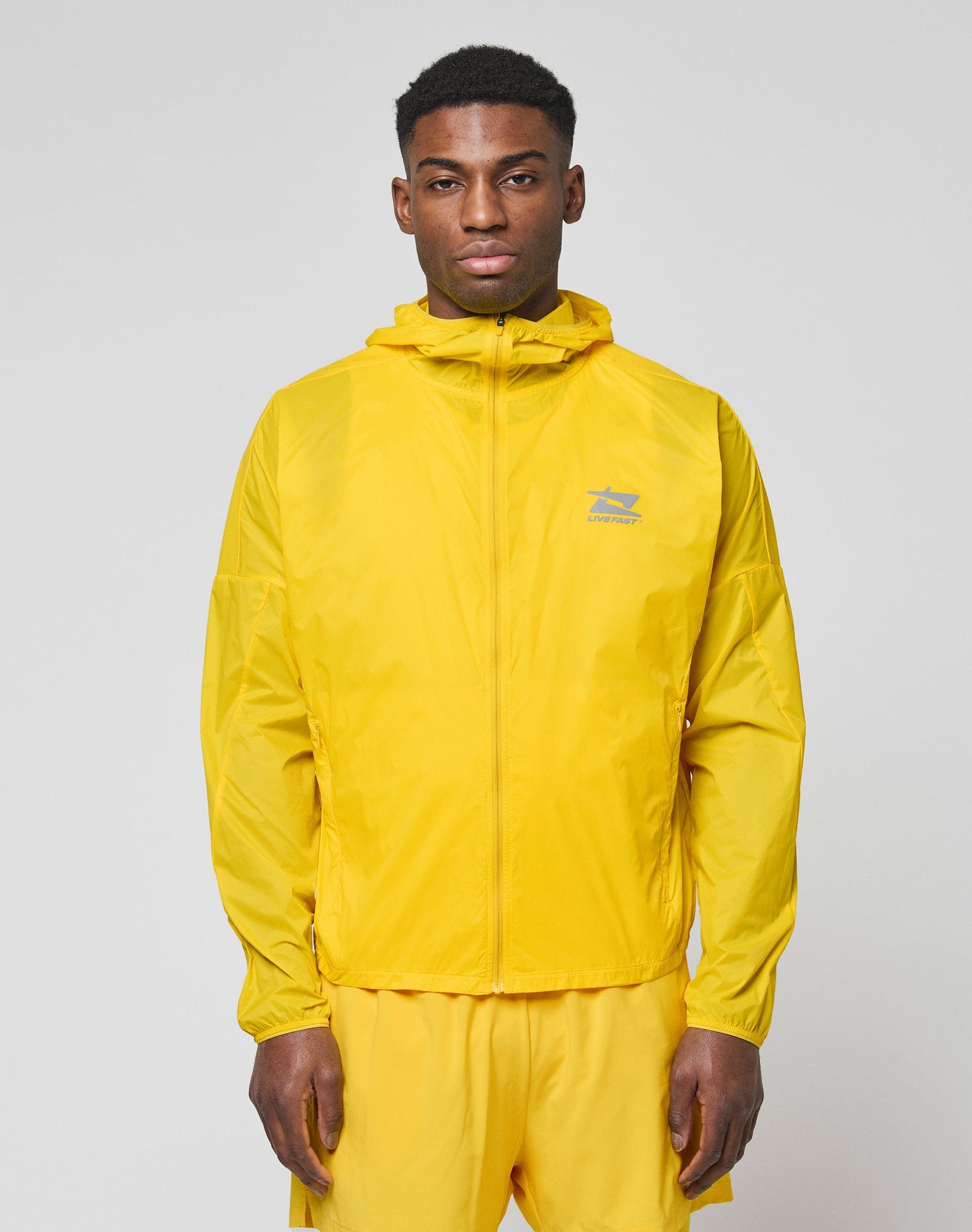A man faces the camera wearing a bright yellow LIVE FAST DIE YOUNG Live Fast Performance Running Jacket with Kapuze mit Visor and matching yellow shorts. The jacket features a small logo on the left chest, set against a plain light gray background.| model