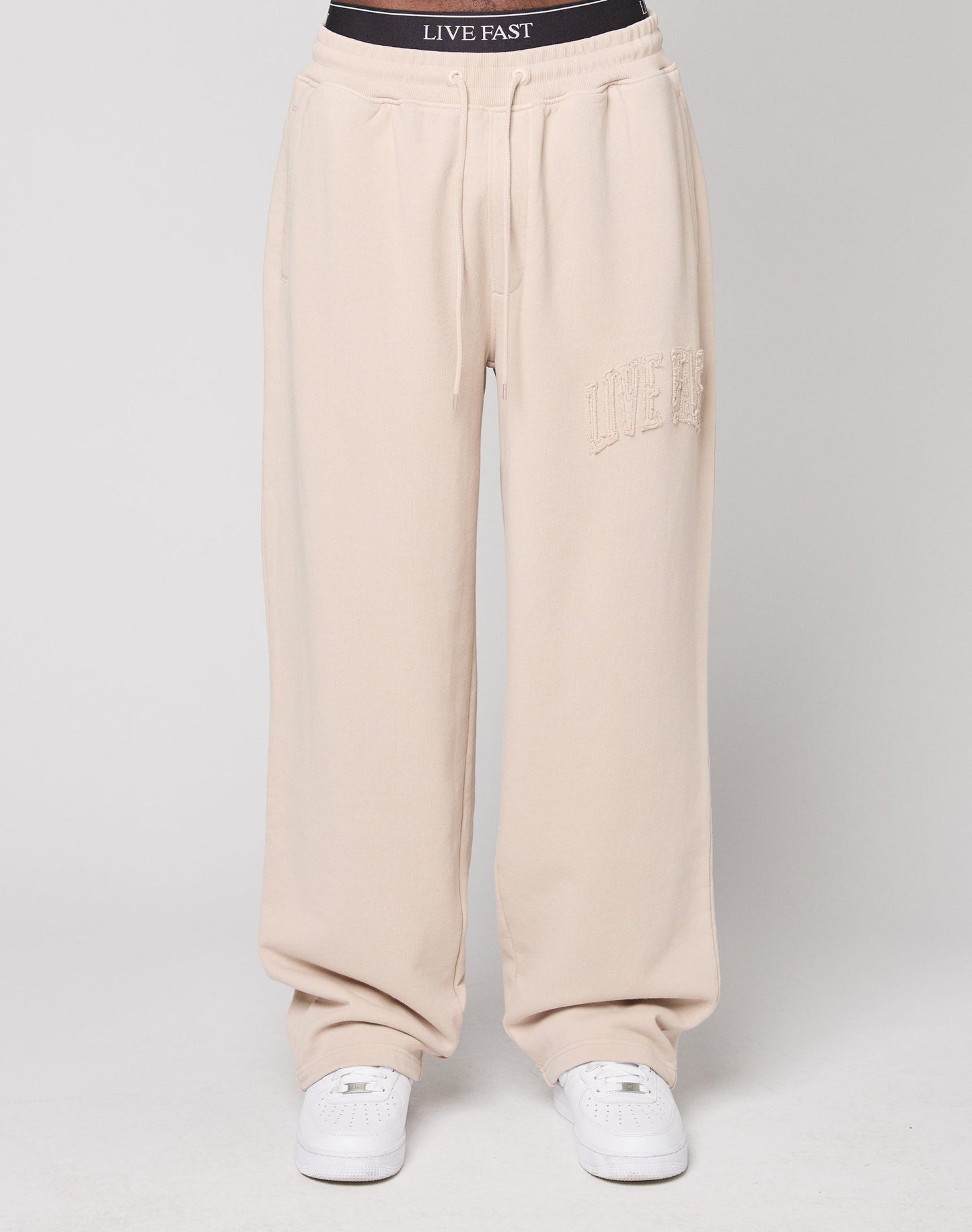 A person wears LIVE FAST DIE YOUNG Ripped College Wide Jogger sweatpants in light beige with LIVE FAST on one thigh, paired with white sneakers and black underwear showing its LIVE FAST branding. The background is plain white.