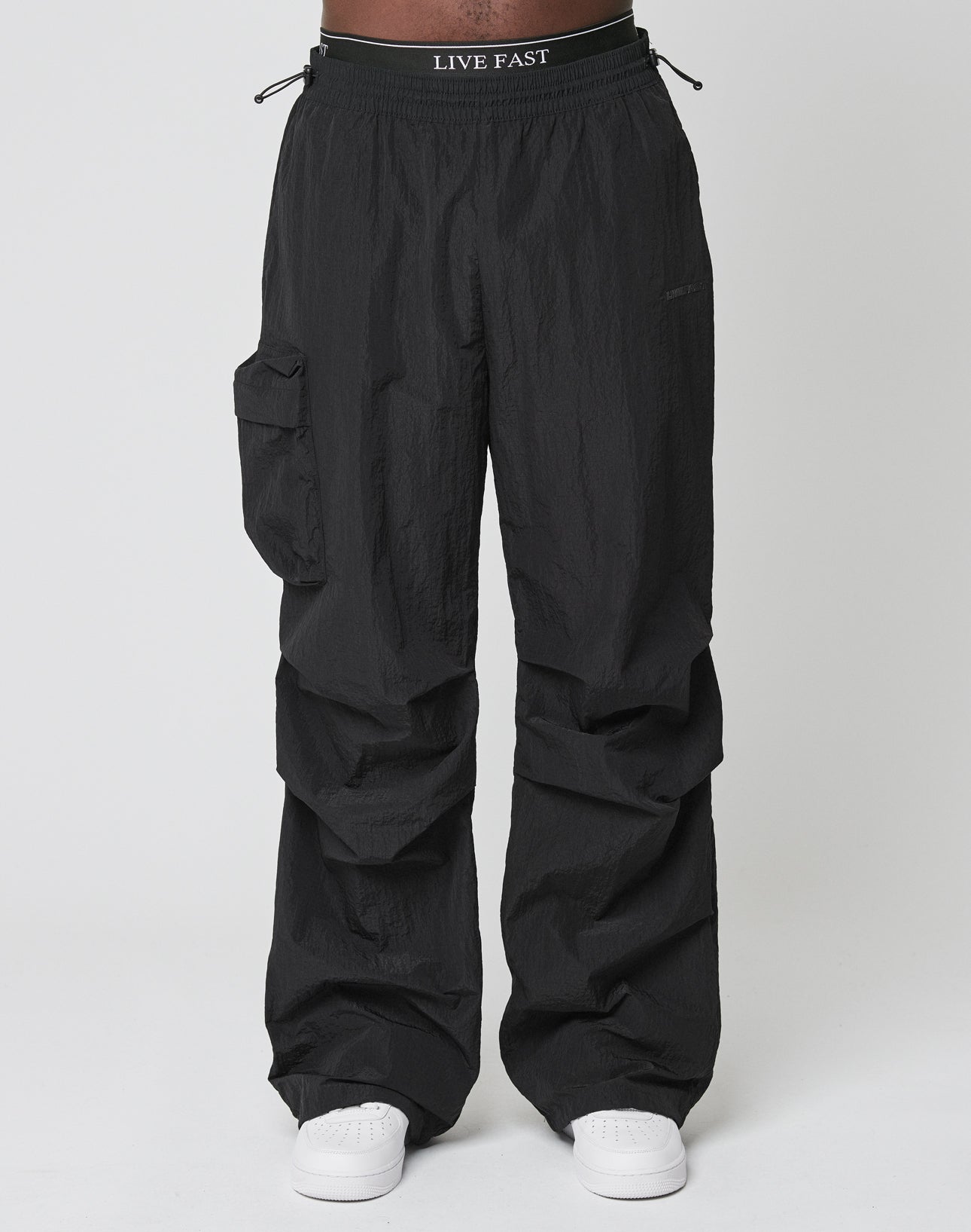 Model wears LIVE FAST DIE YOUNGs Live Fast Nylon Pants—oversized black cargo pants with elastic waistband, cargo pocket, and LIVE FAST print—styled with white sneakers, shot against a plain white backdrop.