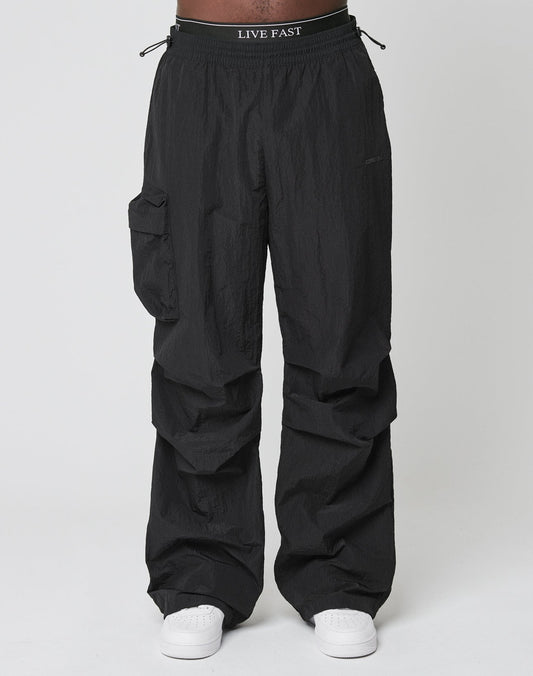 Model wears LIVE FAST DIE YOUNGs Live Fast Nylon Pants—oversized black cargo pants with elastic waistband, cargo pocket, and LIVE FAST print—styled with white sneakers, shot against a plain white backdrop.