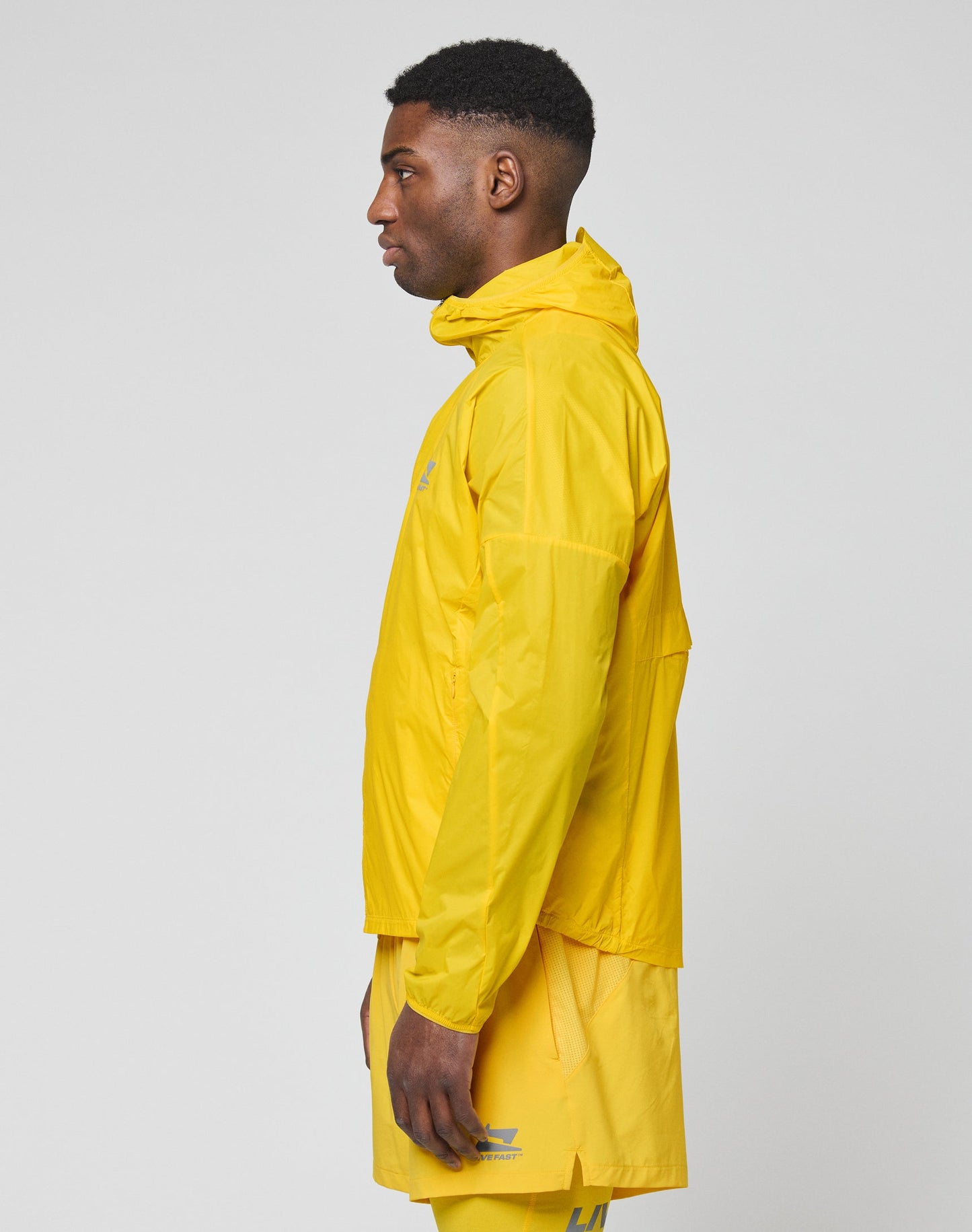 A person models the bright yellow Live Fast Performance Running Jacket by LIVE FAST DIE YOUNG, featuring a visor hood and Regular Fit, standing out against a plain light gray background.