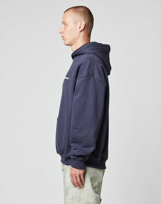 A man in profile faces left, wearing the LIVE FAST DIE YOUNG Live Fast Plain Hooded sweatshirt—oversized, dark blue, soft Baumwolle with front pocket—paired with light pants against a plain gray background.
