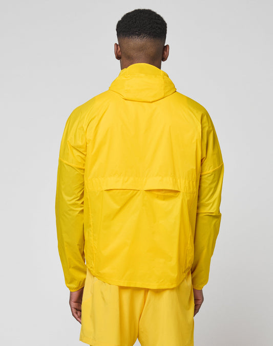 A person wearing the LIVE FAST DIE YOUNG Live Fast Performance Running Jacket in bright yellow with a visor hood and matching yellow shorts stands with their back to a plain light gray background.