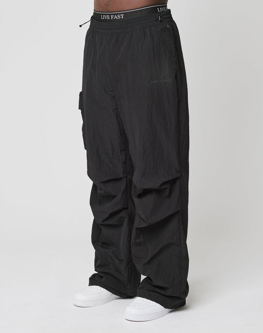 Person wearing Live Fast Nylon Pants by LIVE FAST DIE YOUNG—loose black cargo pants with elastic waistband and cargo pockets, featuring LIVE FAST text, paired with white sneakers against a plain white background.