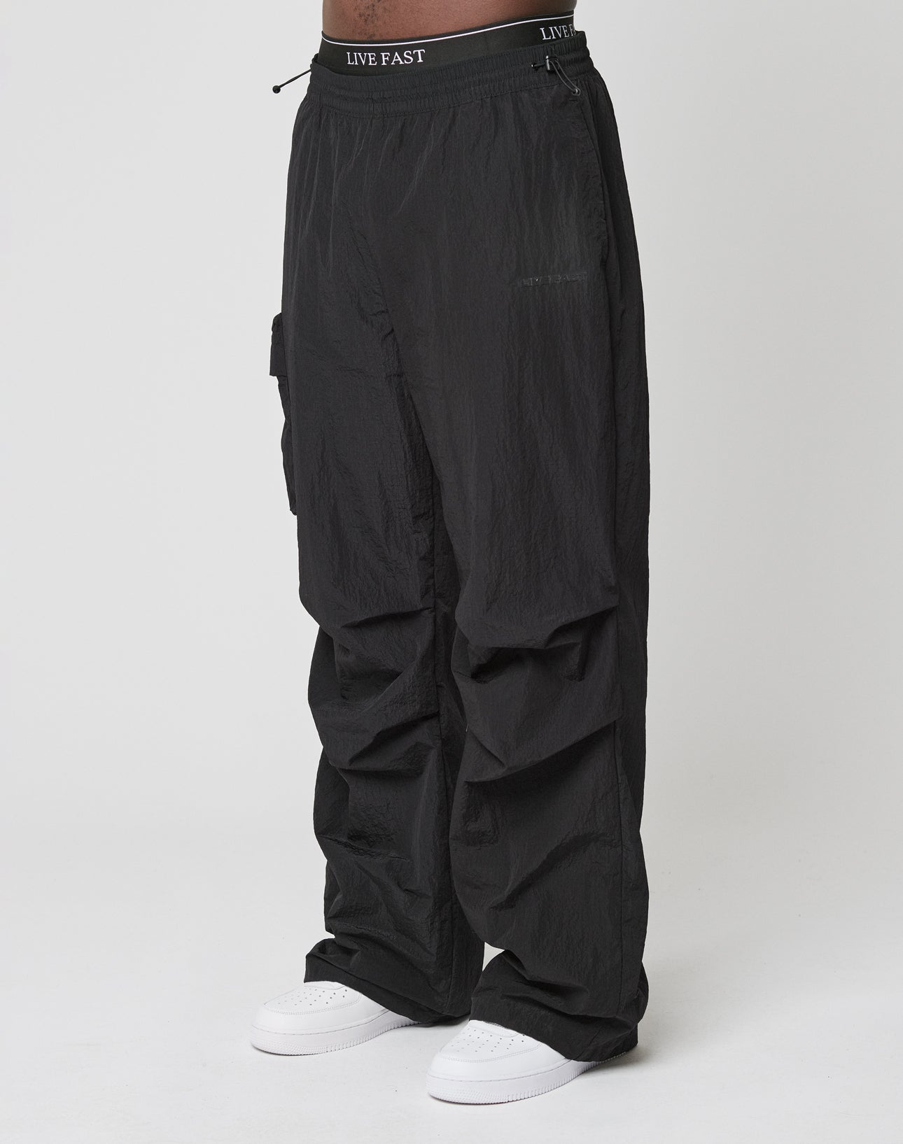 Person wearing Live Fast Nylon Pants by LIVE FAST DIE YOUNG—loose black cargo pants with elastic waistband and cargo pockets, featuring LIVE FAST text, paired with white sneakers against a plain white background.
