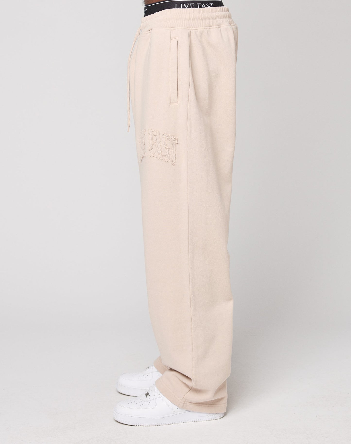 A person is shown in side profile against a plain light background, wearing LIVE FAST DIE YOUNG Ripped College Wide Joggers in beige with textured side lettering and paired with white sneakers.