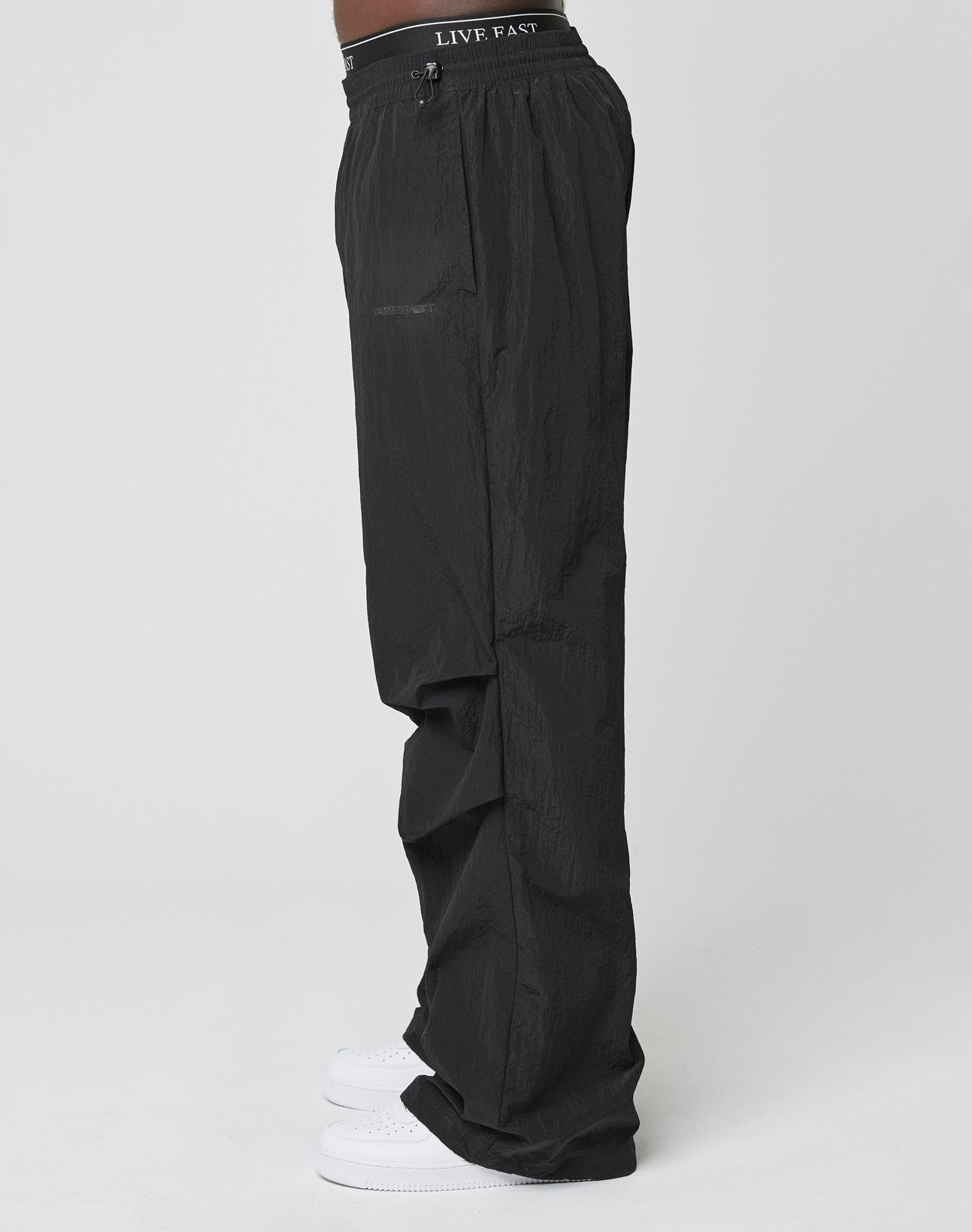 A person in profile wears LIVE FAST DIE YOUNG’s black Live Fast Nylon Pants with a crinkled finish, elastic waistband featuring “LIVE FAST,” and white sneakers, set against a plain white background.
