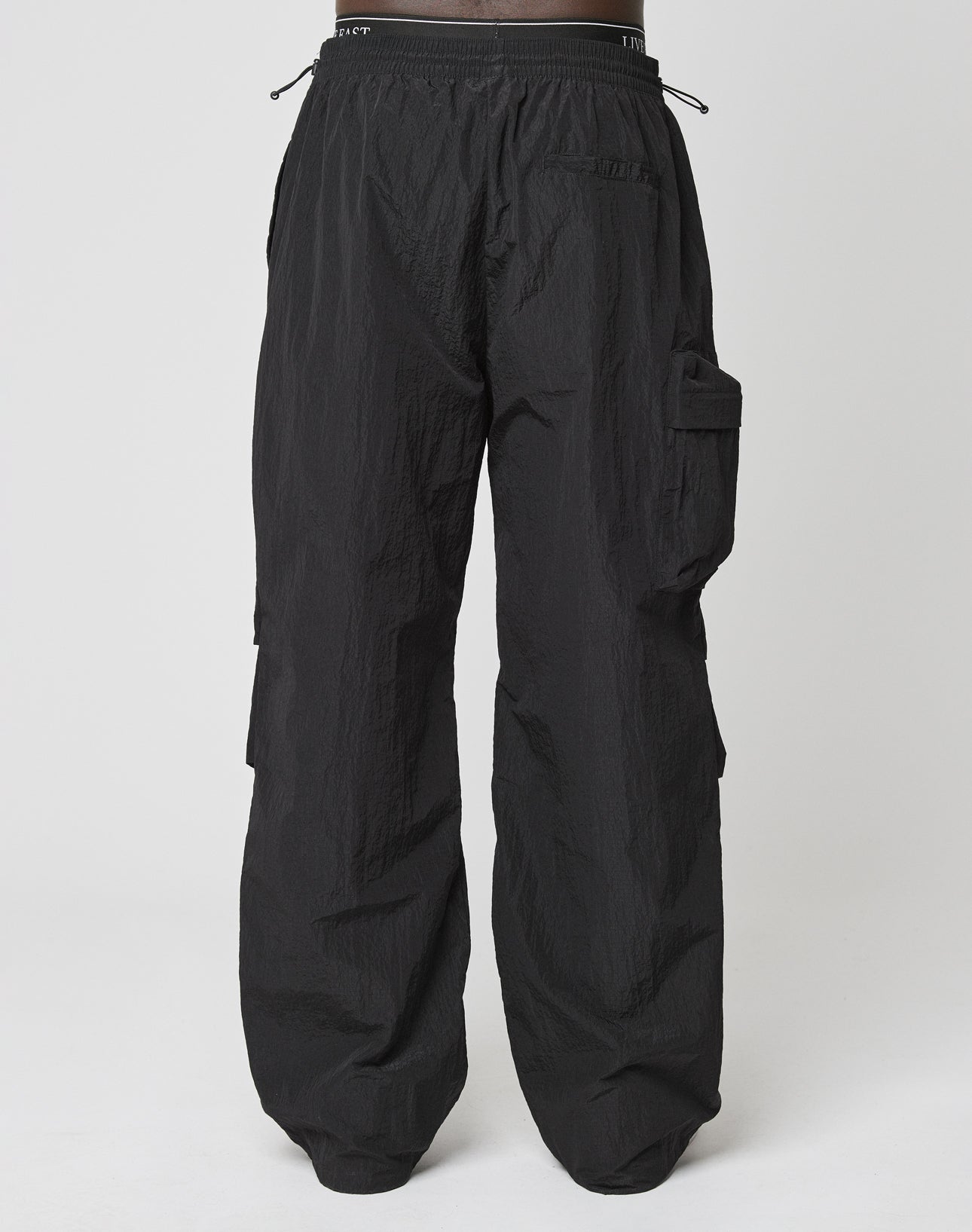 The LIVE FAST DIE YOUNG Live Fast Nylon Pants are black, loose-fitting cargo pants with a crinkled texture, elastic waistband with drawstring, side pockets, and a right-leg cargo pocket in an oversized fit against a white background.