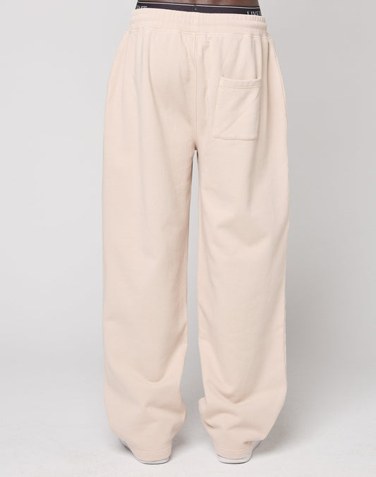 A person wears LIVE FAST DIE YOUNGs Ripped College Wide Jogger, wide fit sweatpants in light beige Baumwolle with an elastic waistband and single front pocket, standing against a plain light gray background.