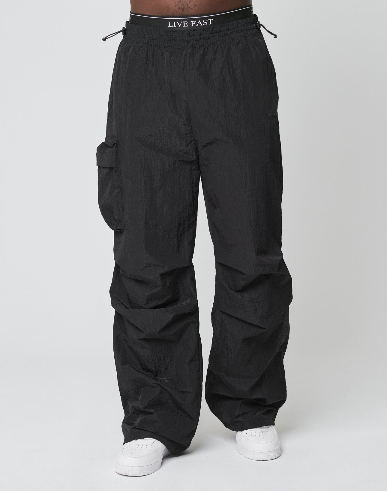 A person wears LIVE FAST DIE YOUNG’s Live Fast Nylon Pants—loose black cargo pants with Cargotasche, Elastikbund, and LIVE FAST waistband—paired with white sneakers against a plain light background.