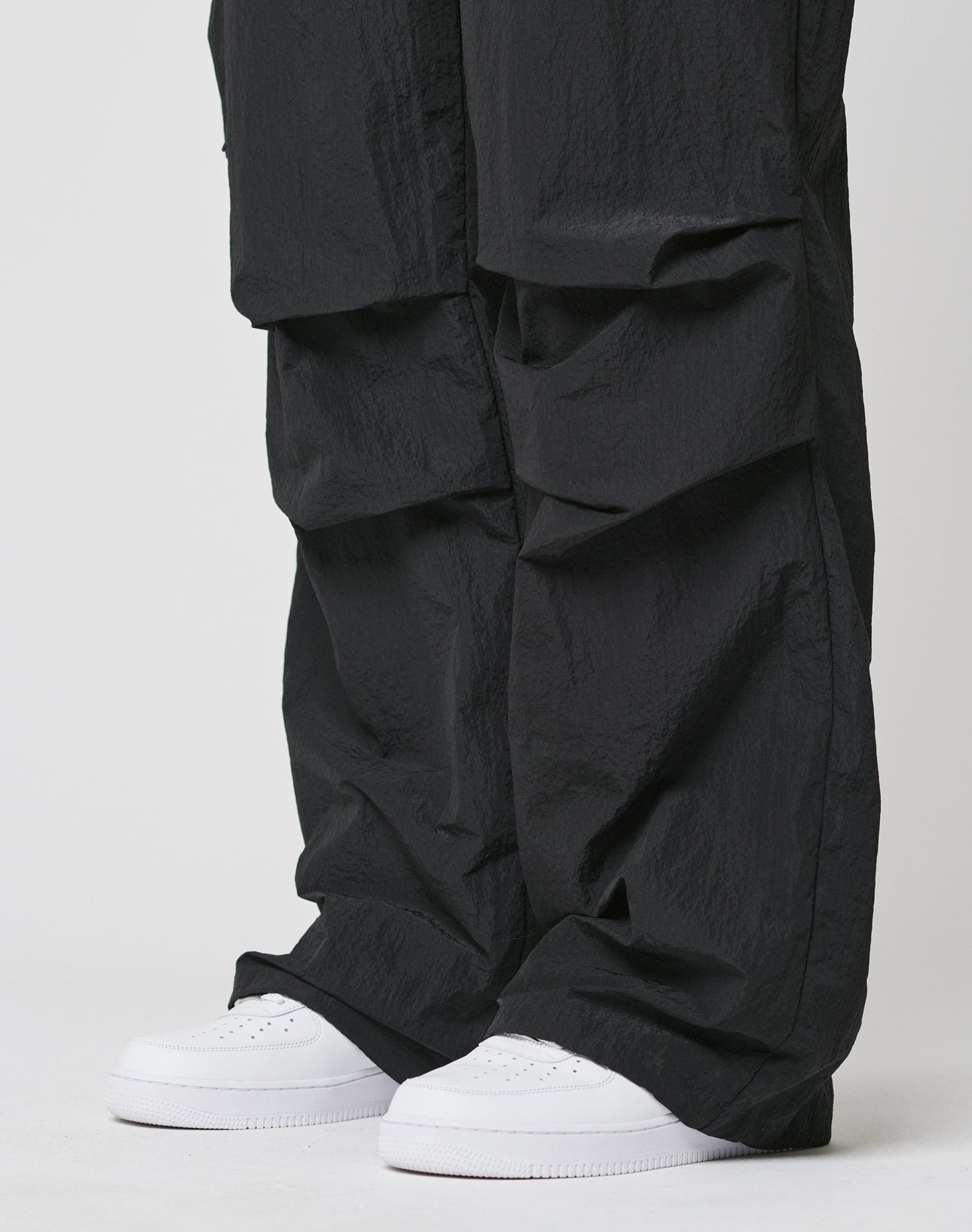 A person wearing LIVE FAST DIE YOUNG Live Fast Nylon Pants in an oversized black fit with elastic waistband, styled over clean white sneakers, stands against a plain white background.