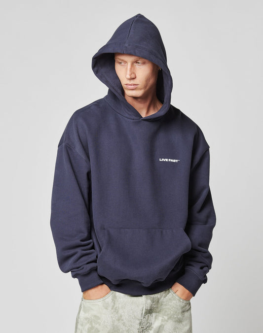A person wears the LIVE FAST DIE YOUNG Live Fast Plain Hooded in dark blue, oversized fit with hood up and hands in the front pocket. Made from soft Baumwolle, it has ‘LIVE FAST’ in small white print on the left chest. The person looks to the side.