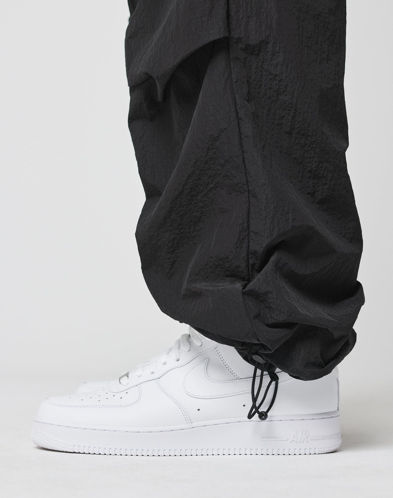 A person wears LIVE FAST DIE YOUNG Live Fast Nylon Pants—loose, black with elastic waistband, ankle drawstrings, and cargo pocket—paired with white Nike Air Force 1s, standing against a plain white background.