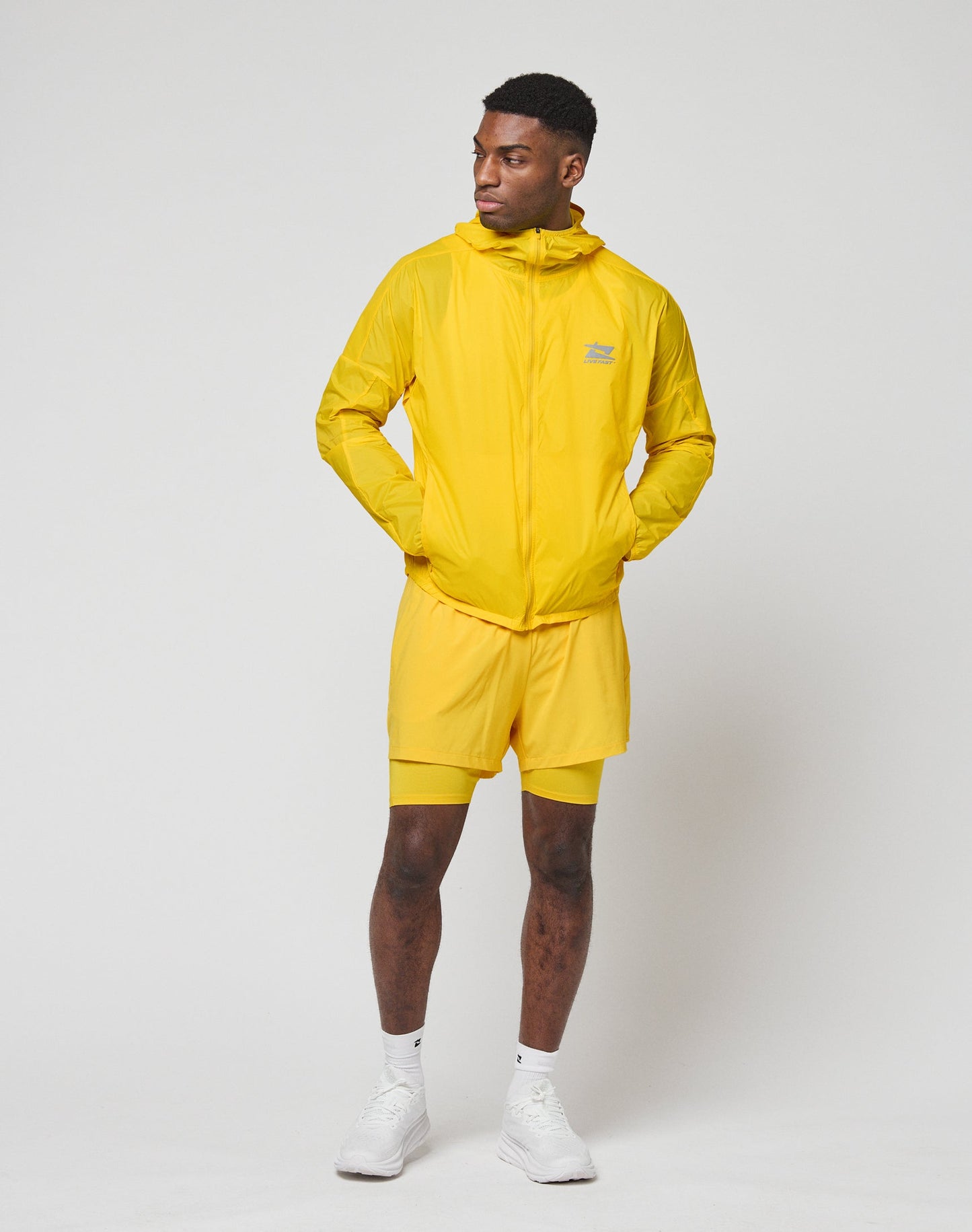 A man stands against a plain background, wearing the LIVE FAST DIE YOUNG Live Fast Performance Running Jacket in bright yellow with Kapuze mit Visor and matching shorts, paired with white socks and athletic shoes. He has a relaxed Regular Fit, hands in pockets.