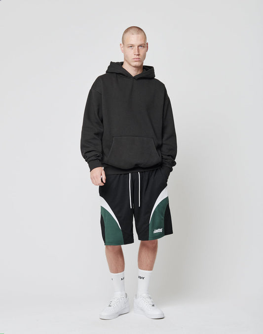 A person with a neutral expression and short hair stands against a plain white background wearing LIVE FAST DIE YOUNG Basic Trackshorts (black/green with white trim, elastic waistband), a black hoodie, white socks, and white sneakers.