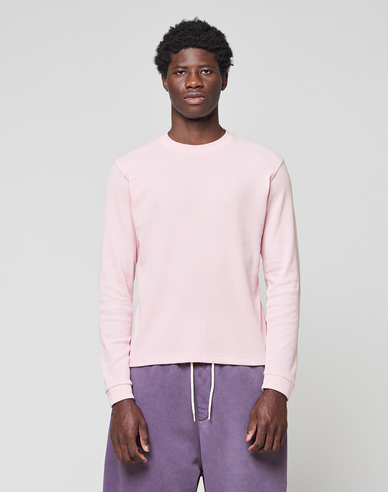 A man stands facing forward, wearing a LIVE FAST DIE YOUNG Basic Waffle Longsleeve in light pink and loose-fitting purple Oversize Fit pants with a white drawstring, set against a plain light gray background. | model