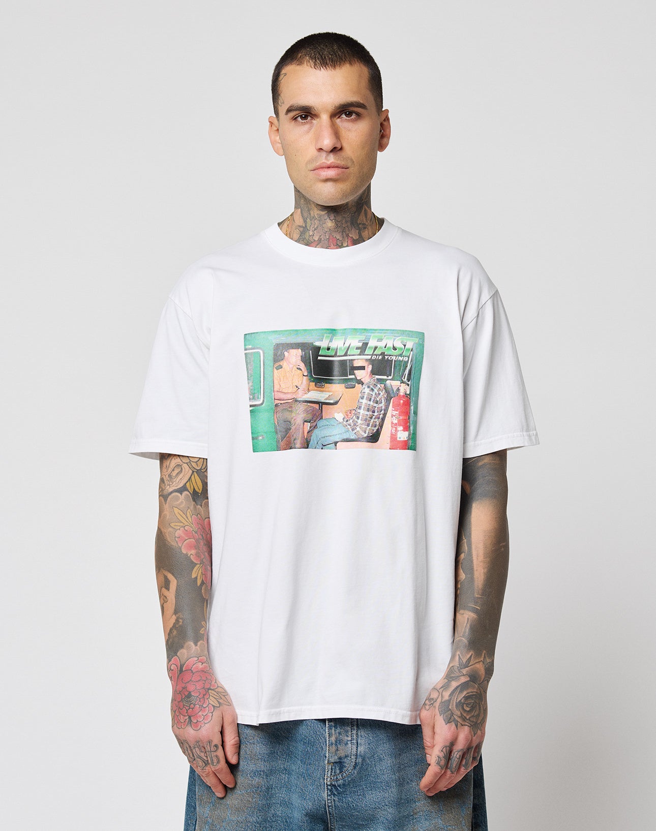 A tattooed man wears the LFDY Sixers Tee from LIVE FAST DIE YOUNG, a white 100% cotton shirt with a colorful two-person car graphic and LIVE FAST text, paired with blue jeans against a plain light background.