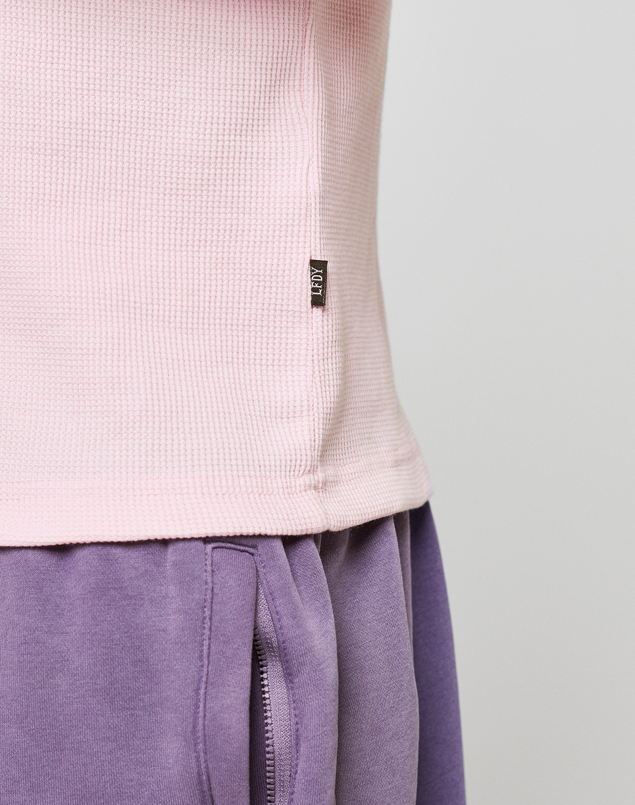 Close-up of a person wearing the Basic Waffle Longsleeve by LIVE FAST DIE YOUNG in light pink with ribbed cuffs and a black tag, paired with high-waisted purple sweatpants featuring oversize fit, visible pocket, and seam detail.