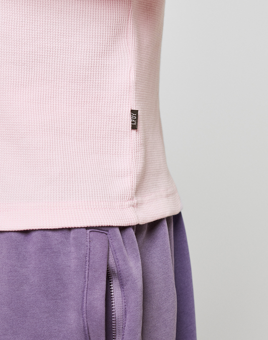 Close-up of a person wearing the Basic Waffle Longsleeve by LIVE FAST DIE YOUNG in light pink with ribbed cuffs and a black tag, paired with high-waisted purple sweatpants featuring oversize fit, visible pocket, and seam detail.