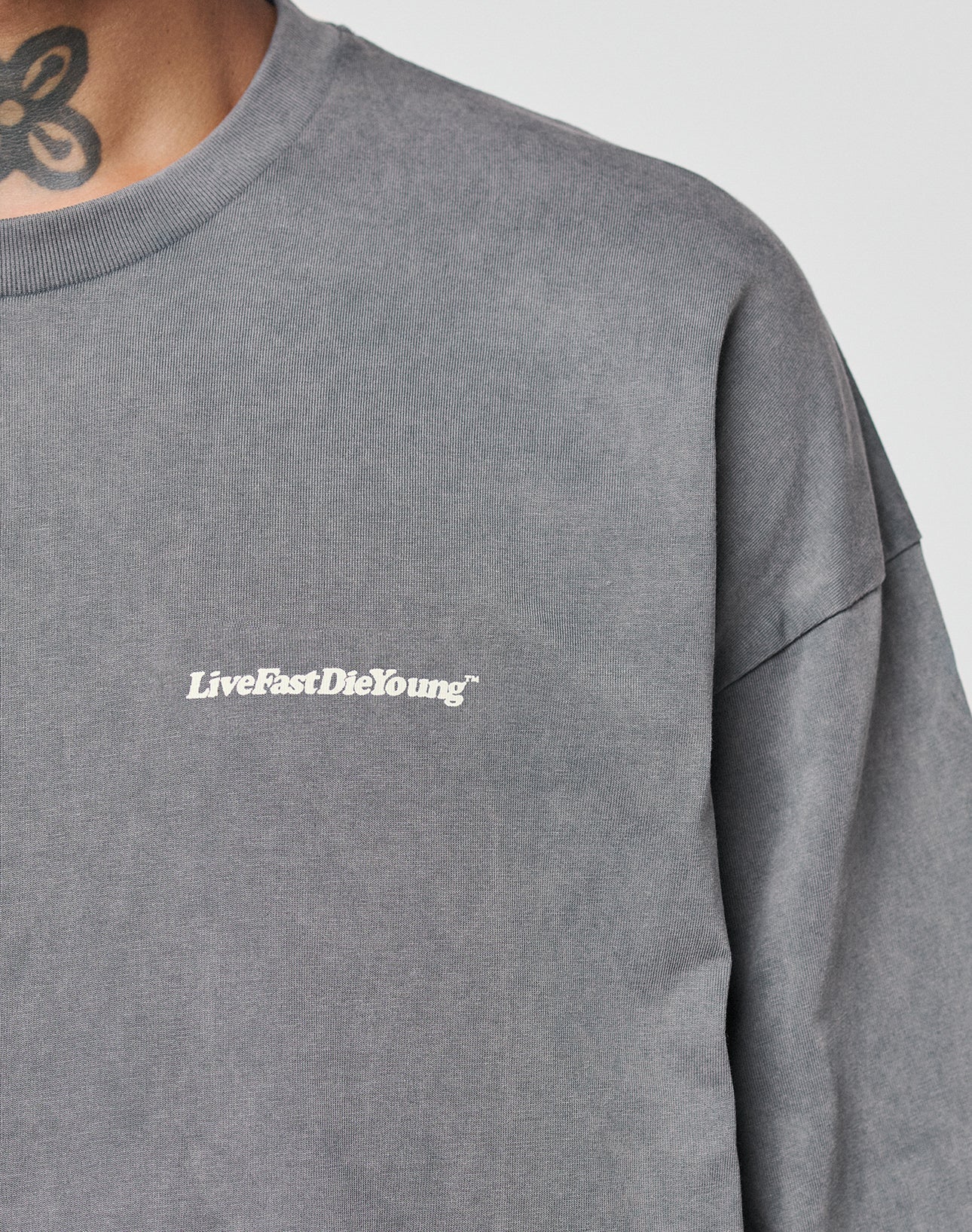 A close-up of a person wearing the LIVE FAST DIE YOUNG Basic Longsleeve, an oversize fit gray sweatshirt with a white LiveFastDieYoung chest logo. Part of a floral neck tattoo is visible above the collar. Made from 100% Baumwolle.