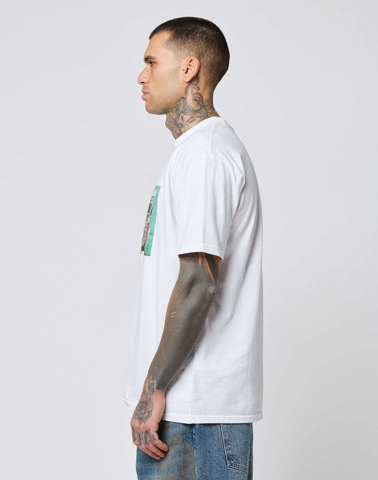 A tattooed man stands in profile facing left, wearing a white LFDY Sixers Tee by LIVE FAST DIE YOUNG made of 100% Baumwolle with a small chest graphic and blue jeans. The background is plain light gray.