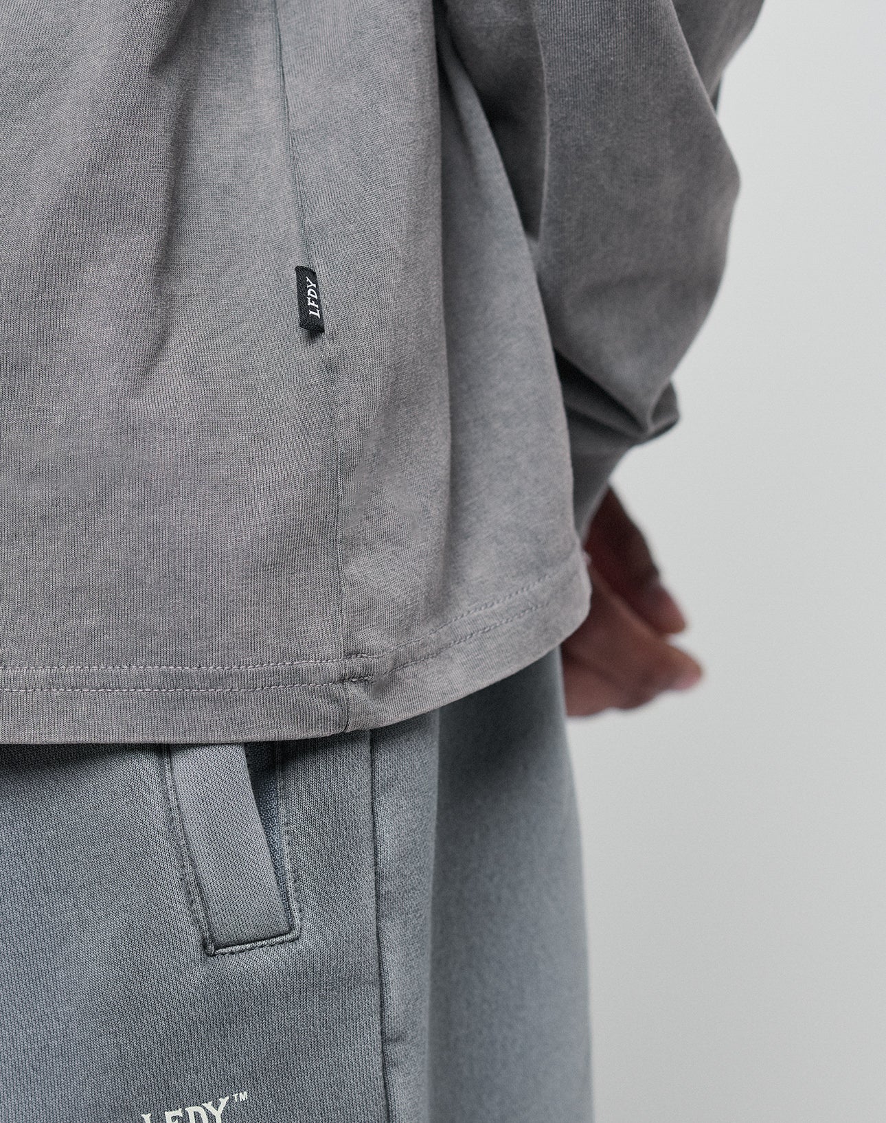 A close-up of a person wearing the LIVE FAST DIE YOUNG Basic Longsleeve and matching gray pants. The 100% Baumwolle shirt has an oversize fit and a small black tag on the side seam, with their hand partially visible.