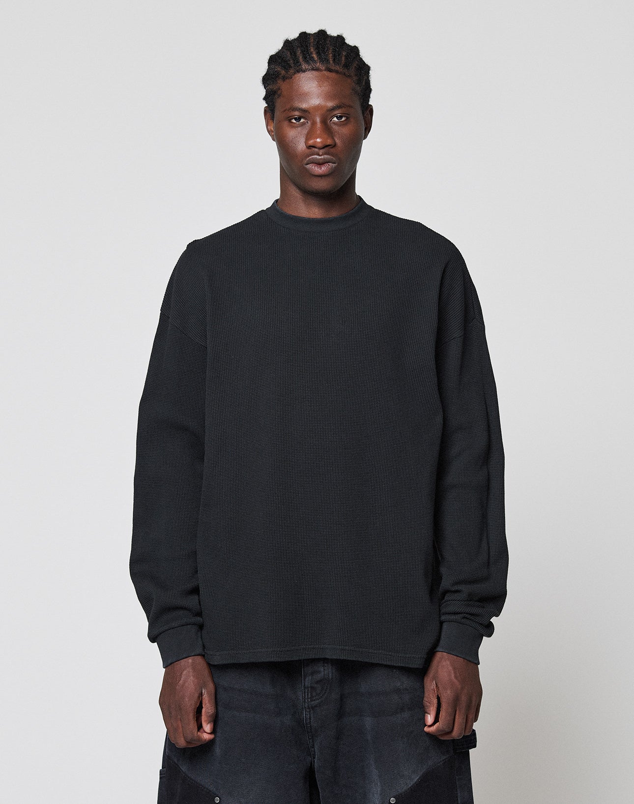 A person with braided hair stands against a plain light gray background, wearing a LIVE FAST DIE YOUNG Blank Waffle Longsleeve in black and black pants, looking directly at the camera with a neutral expression. | model