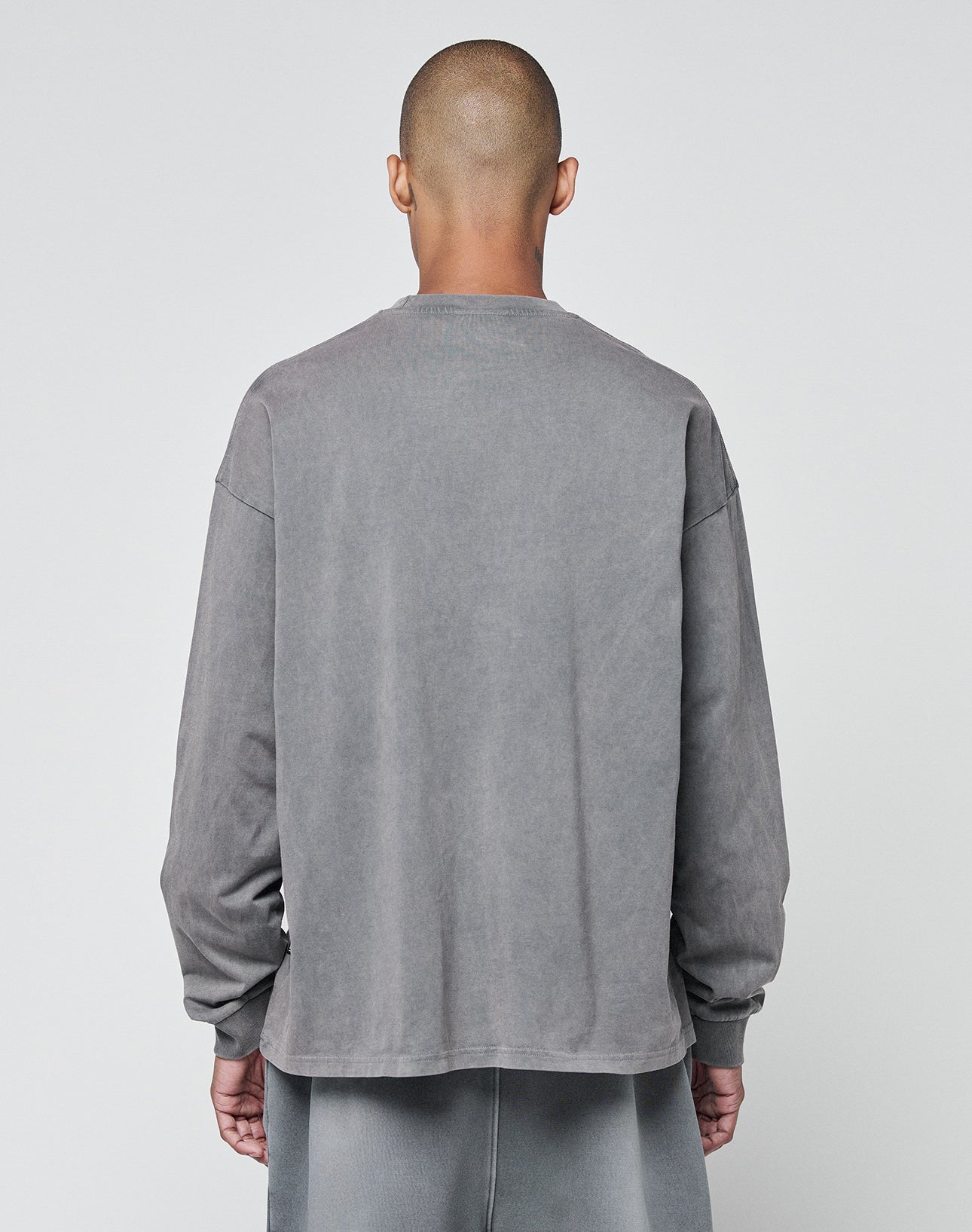 A person with a shaved head stands with their back to the camera, wearing the LIVE FAST DIE YOUNG Basic Longsleeve and matching pants in gray, both made from 100% Baumwolle, against a plain light background.