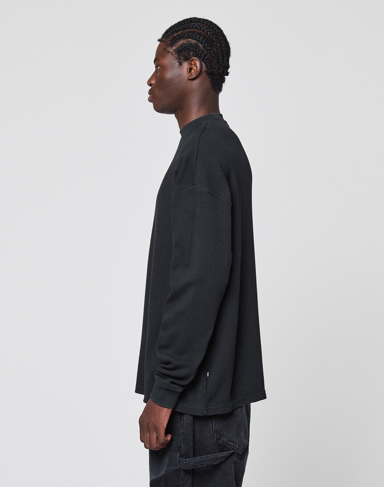 A person with braided hair stands sideways, facing left, wearing the LIVE FAST DIE YOUNG Blank Waffle Longsleeve in black and black pants with Ripp-Bündchen against a plain light gray background.